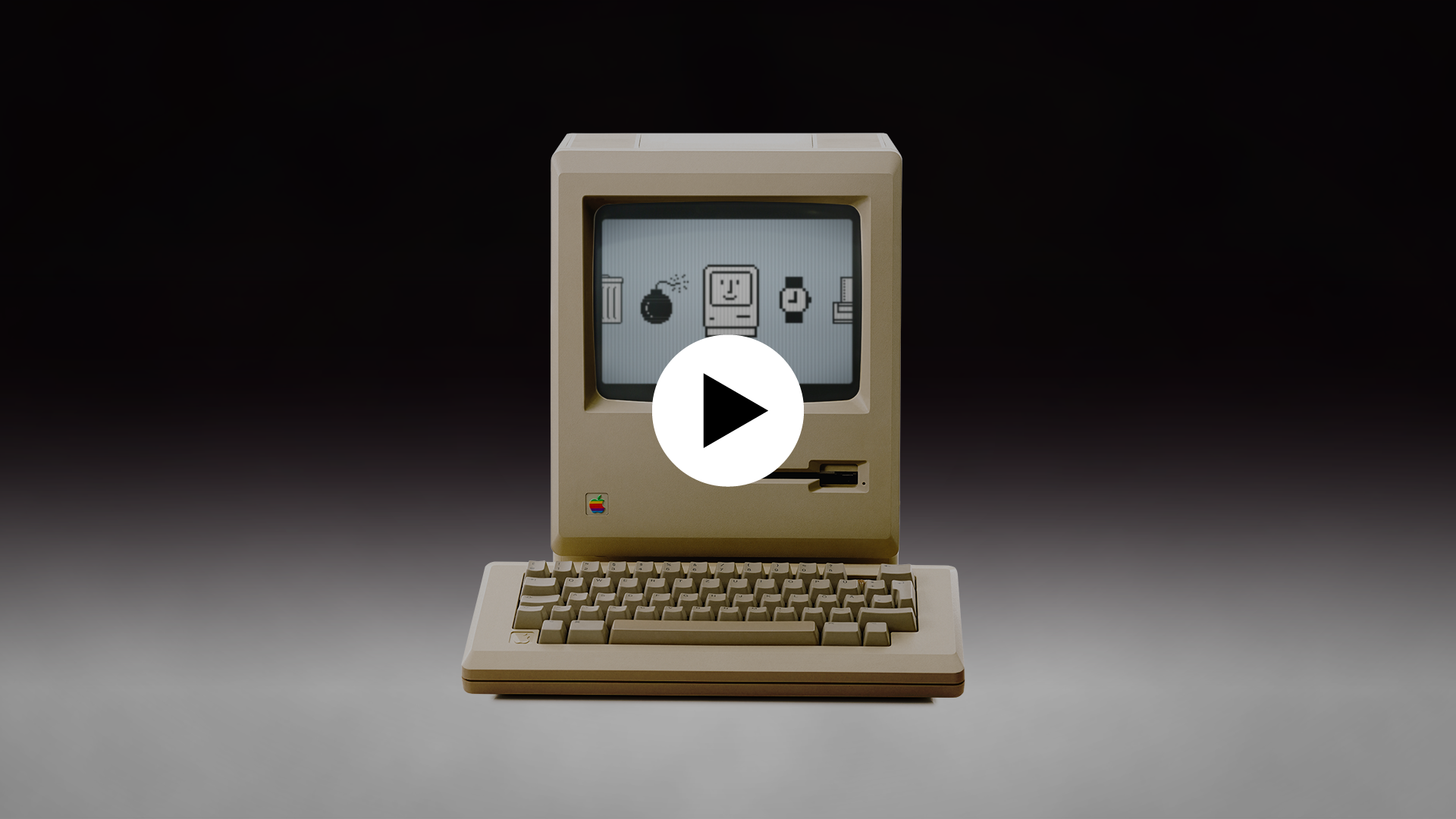 Image du Macintosh original de 1984 sur fond dégradé. Icône play en surimpression indiquant que l'image est cliquable et mène vers un contenu vidéo.